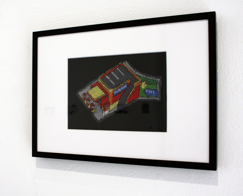 Framed embroidery which shows a petrol station cross stitched on black fabric.