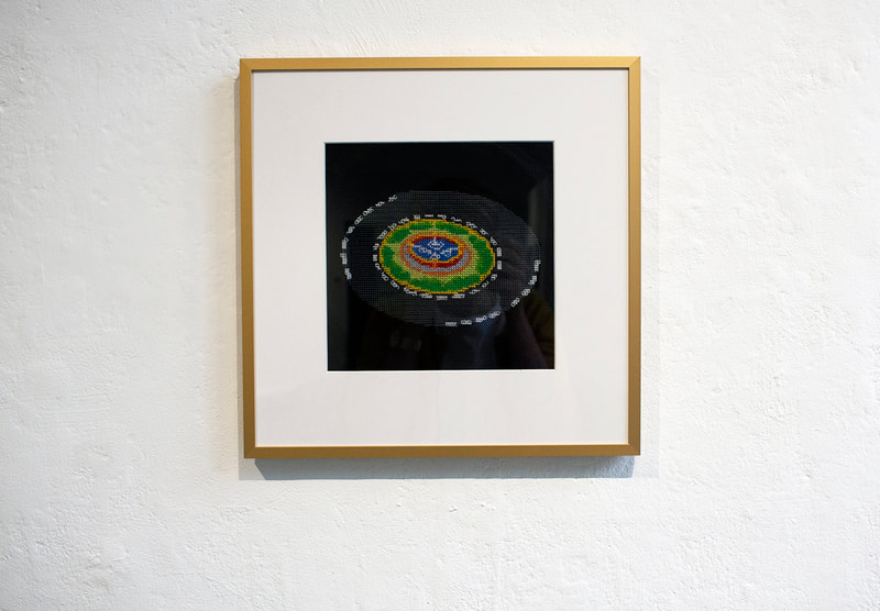 A framed embroidery showing a cross stitched roundabout with a fountain in the middle. The fabric is black, the frame is gold.
